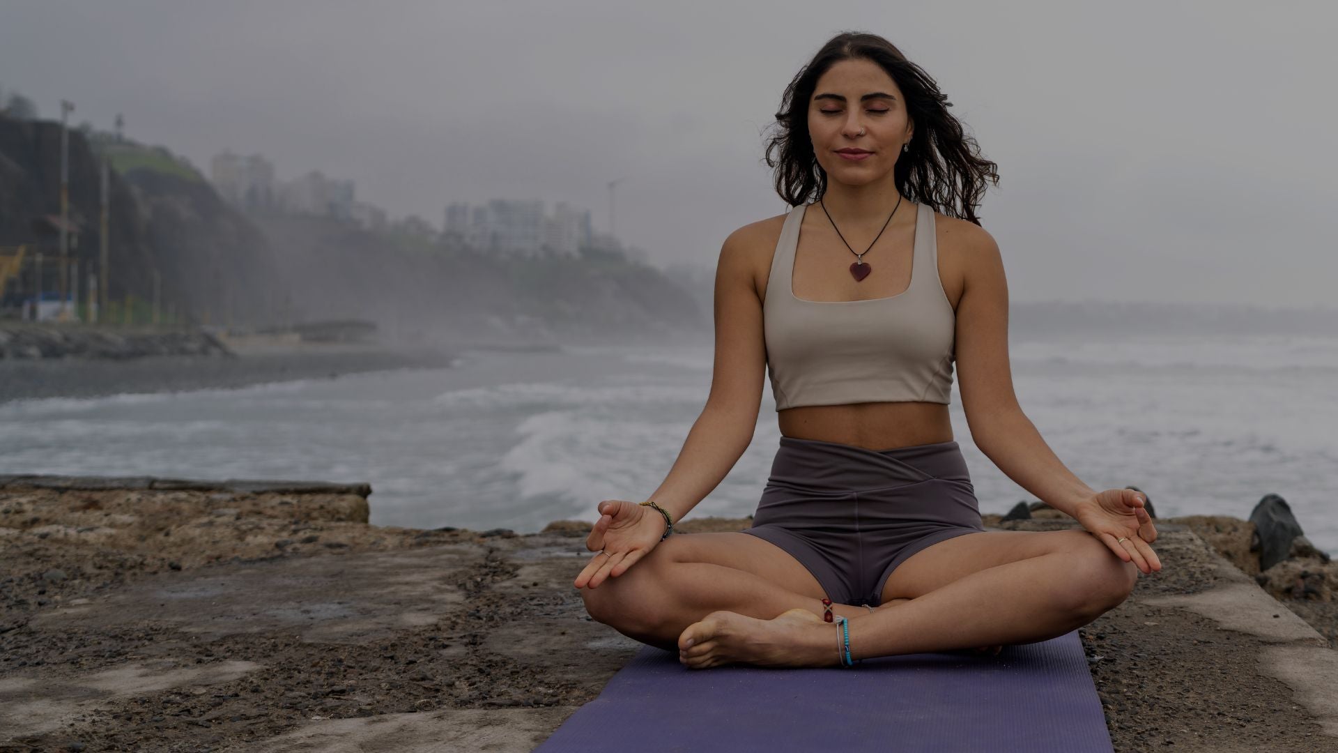 Serene woman sitting in lotus pose meditating on beach rocks with ocean waves in background